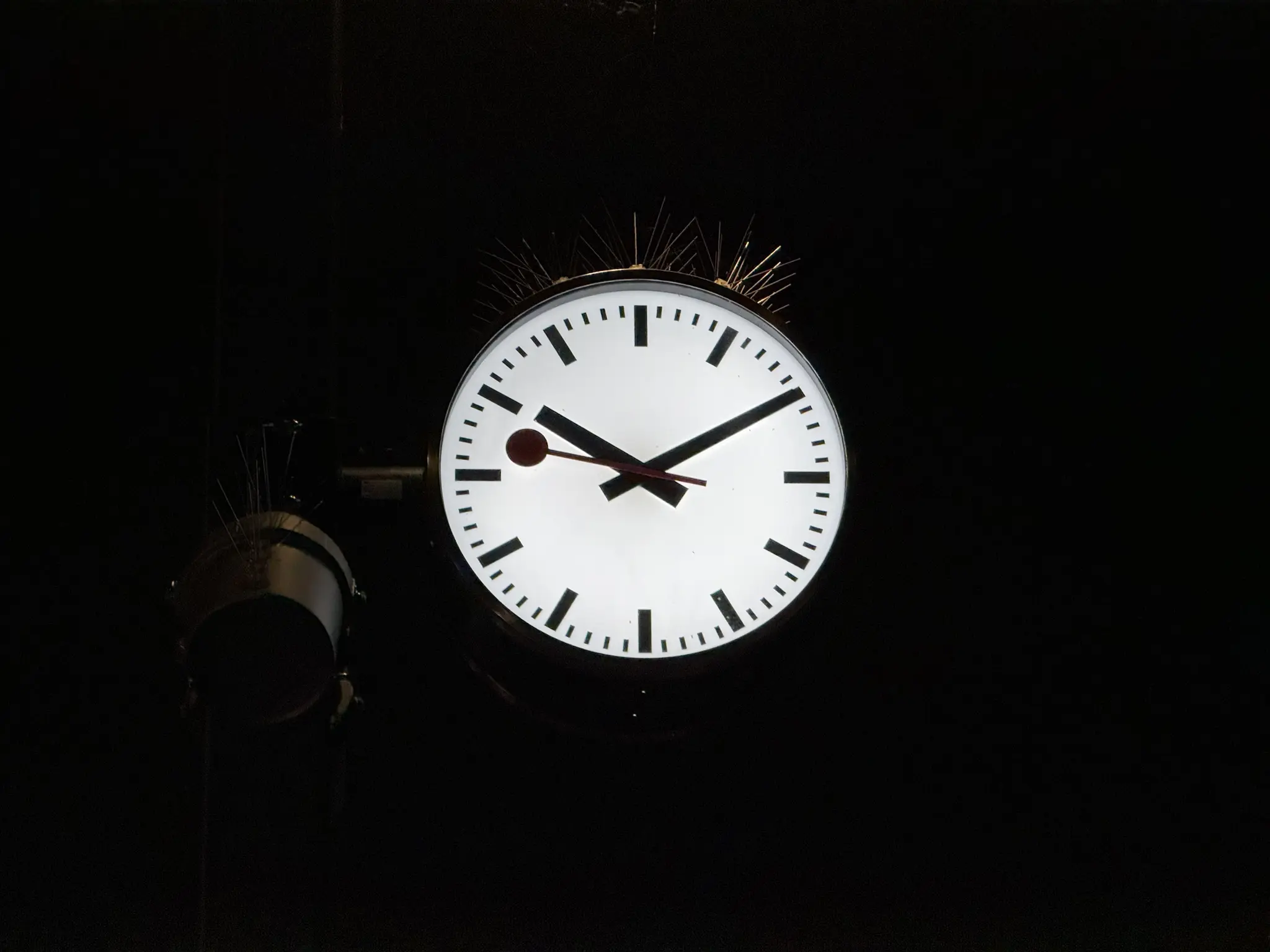 Illuminated SBB clock, only thing clearly visible on the picture, on a post at night displaying the hour: 22:10:47. This and a PA speakers has spikes above them to avoid pigeons to sitting on.